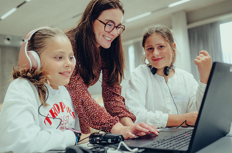 Man sieht zwei Schüler*innen am Laptop mit einer Frau im roten Kleid zwischen ihnen, das linke mädchen schaut in den Bildschirm und hat Kophörer auf