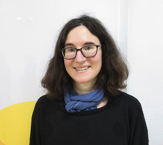 eine weiblich gelesene lächelnde Person, die man von Kopf bis Brust sieht. Sie hat schwarze schulterlange Haare, eine Brille und trägt einen schwarzen Pulli und einen blauen Schal.