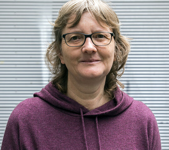 eine weiblich gelesene Person, die von Kopf bis Brust zusehen ist. Sie lächelt in die Kamera, hat graues wildes Haar mit einem kleinen Pony, trägt eine Brille und ein rosarotes Shirt.