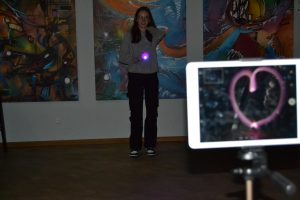 Im Vordergrund sieht man ein iPad auf dem mit einer Langzeitbelichtung gerade ein Bild von einem Herzen gemacht wurde. Im Hintergrund steht eine Teilnehmerin mit einer pink leuchtenden Taschenlampe in der Hand, die wohl gerade das Herz gemalt hat.