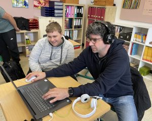 Ein Medienreferent und ein Teilnehmer arbeiten mit Kopfhörern am Laptop. 