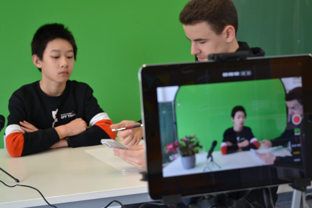 Ein Tablet Bildschirm zeigt die Videoaufnahme zwei junger Menschen, die an einem Tisch sitzen mit dem Blick auf ein Papier und Smartphone.