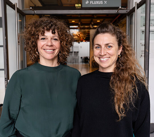 Zwei Frauen mit braunen locken lächeln in die Kamera.
