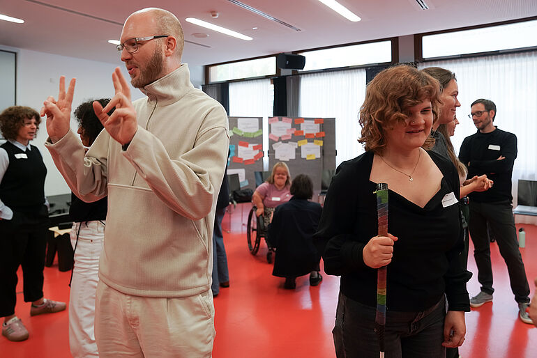 Zwei Menschen sind im Vordergrund zu sehen, links ein Mann mit Brille welcher seine Hände vor sich hält, rechts eine Frau mit Blindenstock.