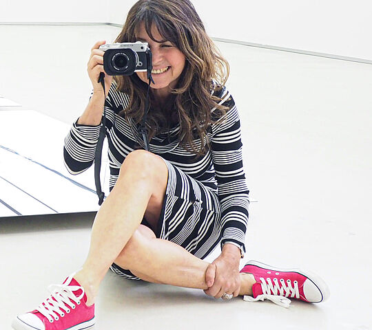 eine braunhaarige Frau, die auf dem Boden sitzt und mit einer Kamera etwas fotografiert. Sie trägt ein schwarz weiß gestreiftes Kleid und dazu pinke Converse.