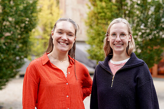 Zwei Frauen lächeln in die Kamera, die rechte Frau hat eine Brille an.