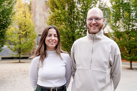 Man sieht eine Frau links, mit langen Haaren, rechts von ihr steht ein Mann mit Brille und ohne Haare. Beide Personen lächeln in die Kamera