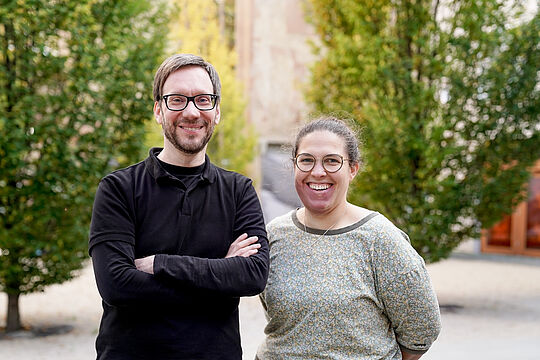 Auf dem Bild ist links ein Mann zu sehen, und rechts eine Frau. Beide lächeln in die Kamera und haben eine Brille auf.